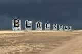 The letters B L A C K A L L on a sign in front of a dark sky full with storm clouds