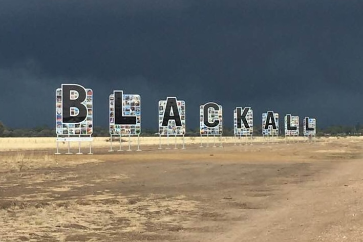 The letters B L A C K A L L on a sign in front of a dark sky full with storm clouds