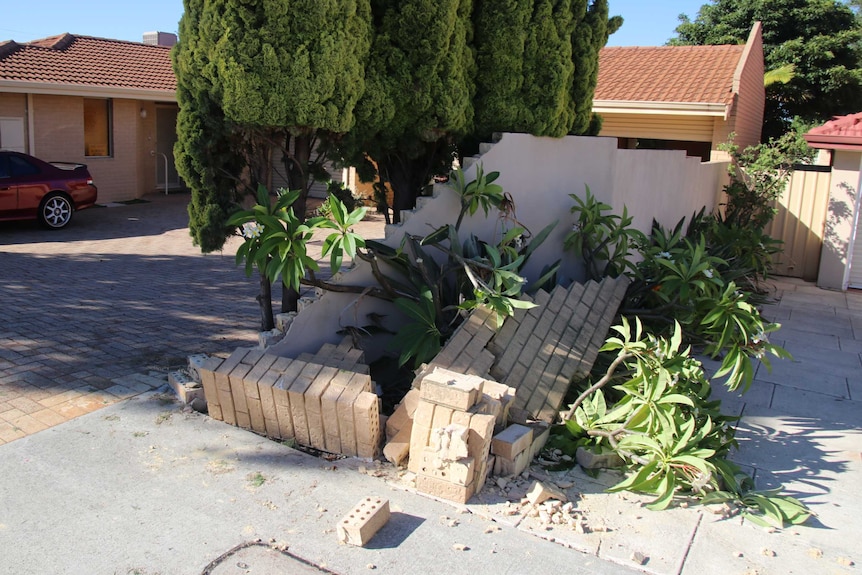 A crumbled wall next to a driveway outside a house, with trees alongside it.