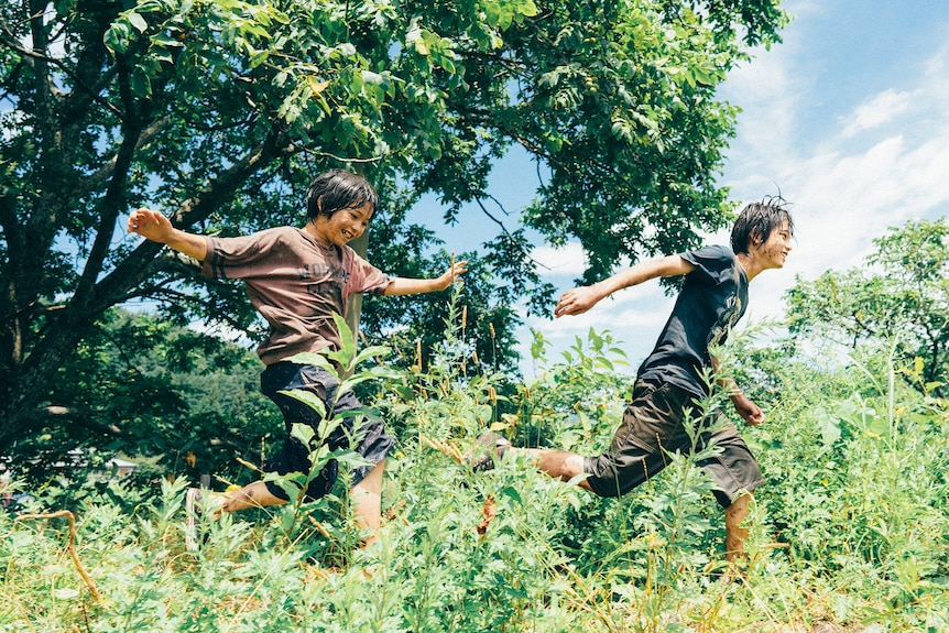 Two young people run through a bushy green area.