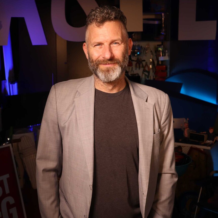 man with grey beard wearing suit standing in dark on TV set only he is illuminated