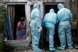 A woman with a cloth mask and purple sari watches three medical workers in blue medical suits enter a doorway.