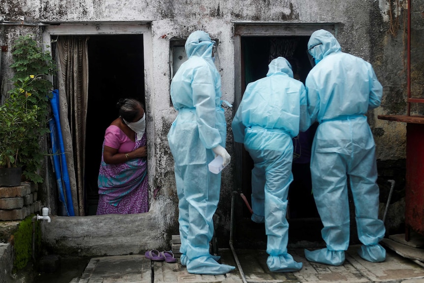 A woman with a cloth mask and purple sari watches three medical workers in blue medical suits enter a doorway.