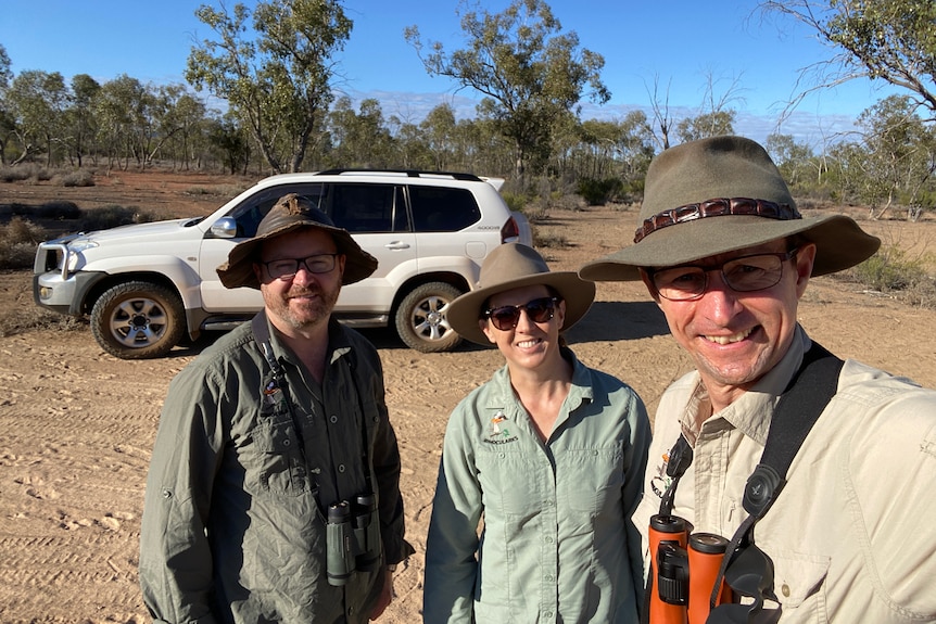 Two men and and a woman stand in long sleeve shirts and broad hats, in a dry bush area, holding binoculars.