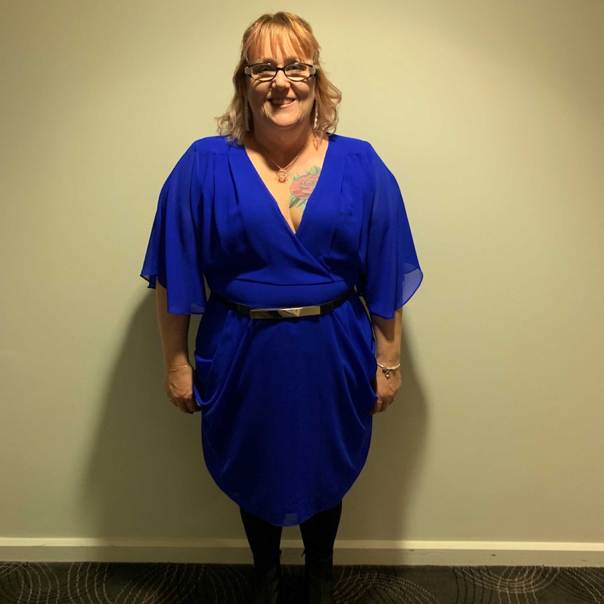 A woman with blonde hair and glasses wearing a blue dress and black boots stands in front of a wall smiling at the camera.