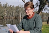A woman sits at a riverside table and holds a piece of paper