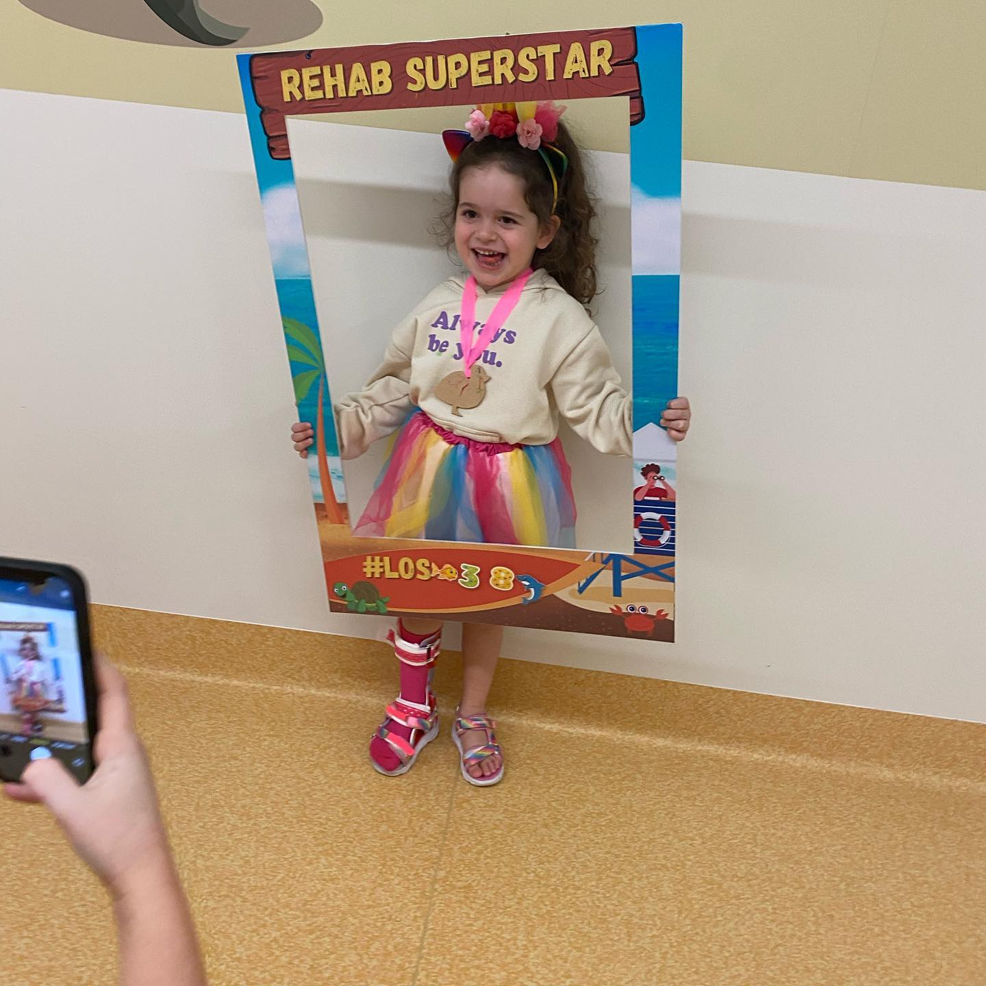 Little girl, Arlo wearing a bright outfit, smiling, holding a sign that says "Rehab Superstar". 