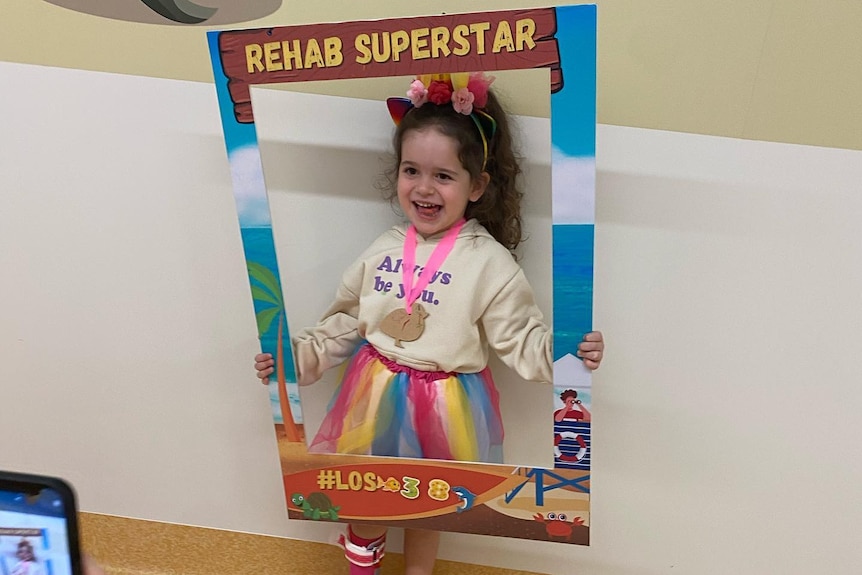 Little girl, Arlo wearing a bright outfit, smiling, holding a sign that says "Rehab Superstar". 