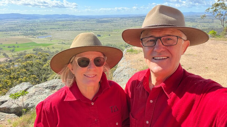 A woman and man in red shirts and broad-brimmed hats smile to camera, with a valley of green farmland in the background.