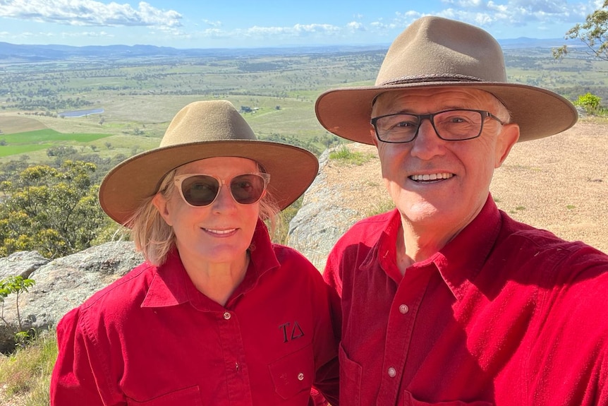 A woman and man in red shirts and broad-brimmed hats smile to camera, with a valley of green farmland in the background.