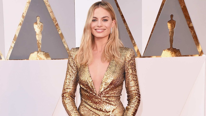 Margot Robbie in a figure hugging, shiny gold full length gown on the red carpet.