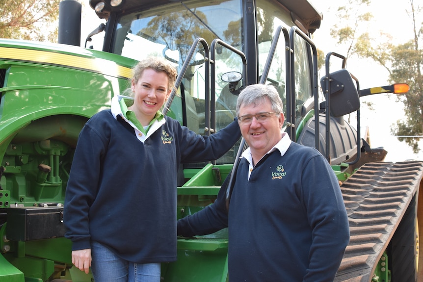 A blonde woman, Dr. Gunn, stands next to a large green tractor with a gray-haired man wearing glasses, Mr. Gladigau.
