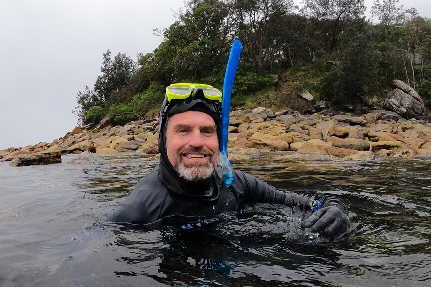 A middle-aged man with a grey beard, wearing a wetsuit, goggles and snorkel, emerges from the water, smiling