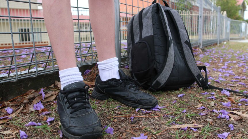 A person stands against a wire fence. They wear black school shoes, white socks and a backpack is seen to the right.