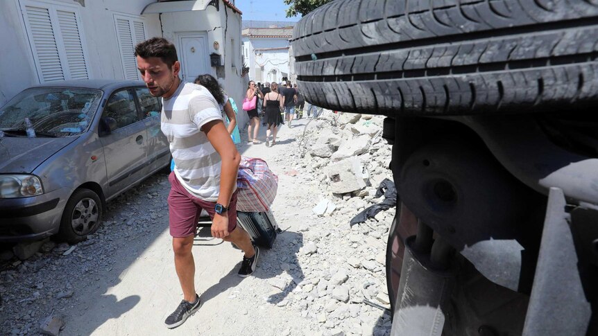 Tourists walk with their suitcases past rubble and an overturned car.