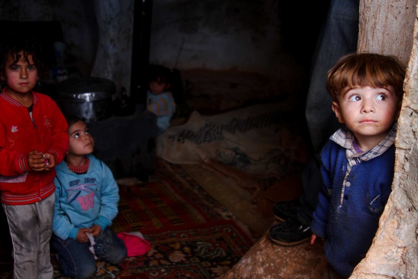 You look down into what appears to be a rock cavern, with children huddled inside. A boy at the entrance stares into the camera.