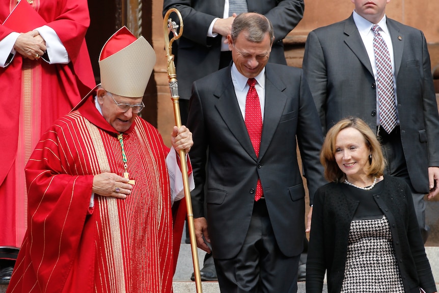 John Roberts walks down church steps with a priest holding a staff and a woman in a black blazer