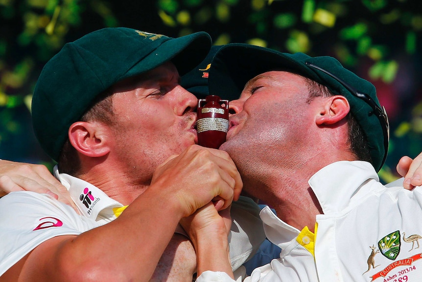 Two men in baggy green hats kiss either side of a small urn