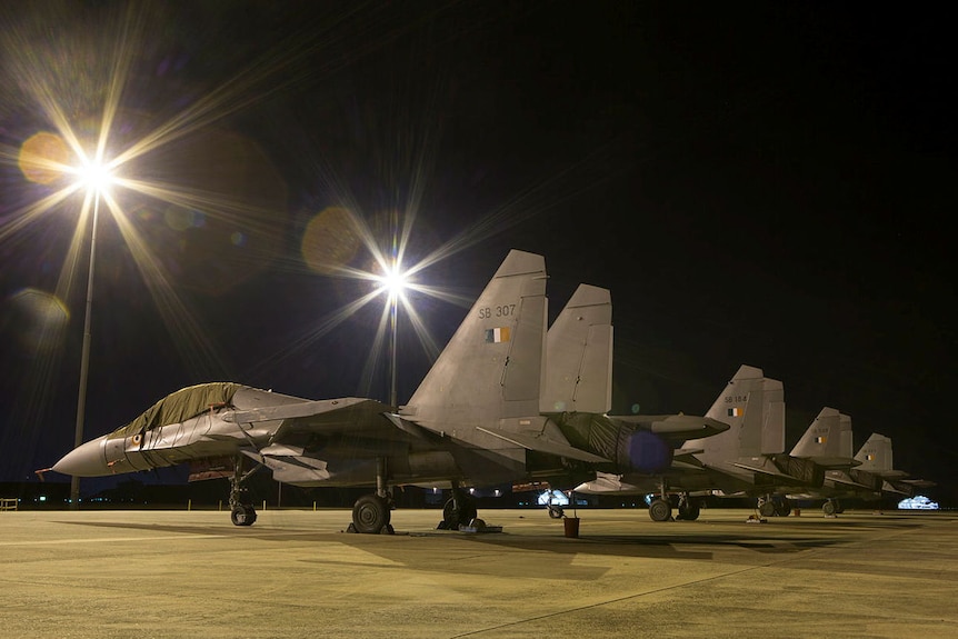 A row of grey, sleek, small military aircraft parked on a tarmac at night, with light flare above some of them.
