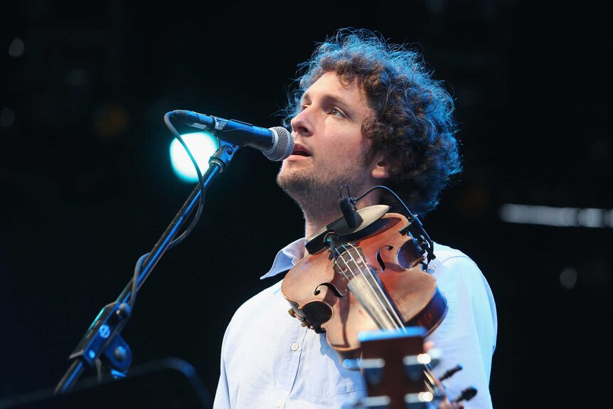 Folk musician Sam Amidon sings into a microphone with a violin on his shoulder.