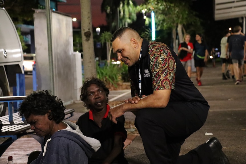 Shannon Brahim kneels down on the street and talks to people at a bus stop.