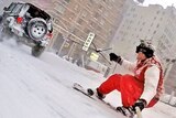 Two men snowboard on the snowy streets of New York City and are towed along by a black car.