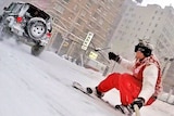 Two men snowboard on the snowy streets of New York City and are towed along by a black car.