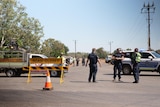 Three police stand next to a road closure sign and witches hat in front of a stretch of bitumen