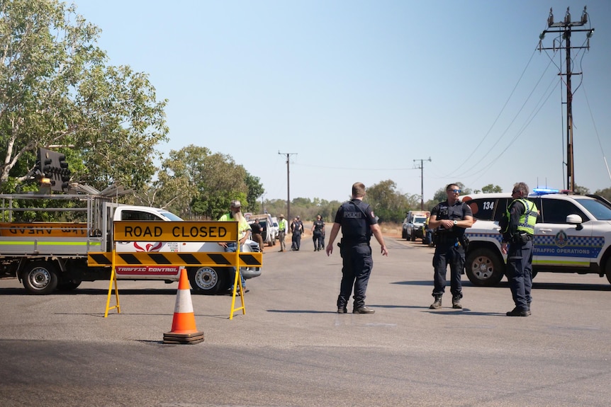 Three police stand next to a road closure sign and witches hat in front of a stretch of bitumen