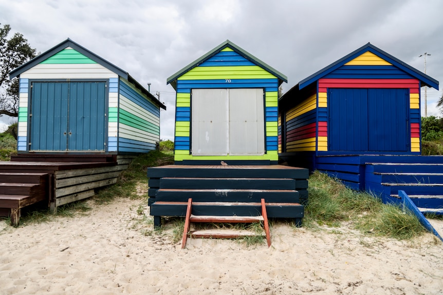 Three boatsheds next to each other painted in bright vibrant colours or blue green orange and red. 