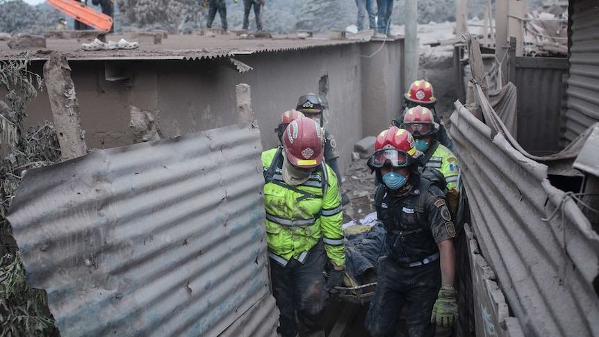 Firefighters carry a body on a stretcher from a damaged building covered in ash.