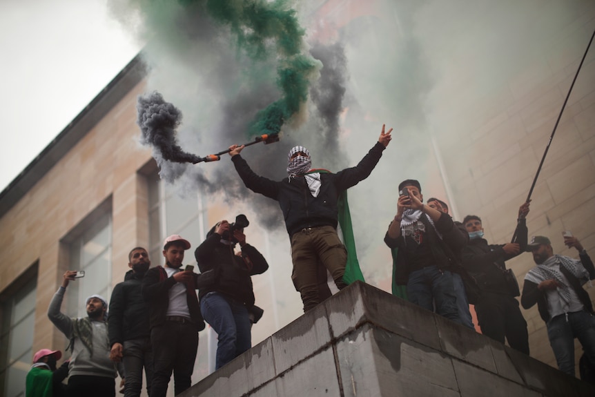 A man holds a smoke flares as he shouts slogans while standing on top of a building with other people.