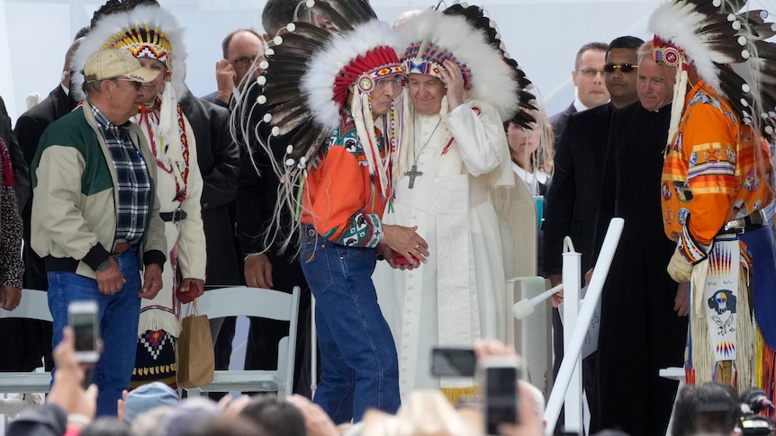A man in traditional dress stands partly in front of Pope Francis who is wearing robes and a traditional Indigenous head piece. 