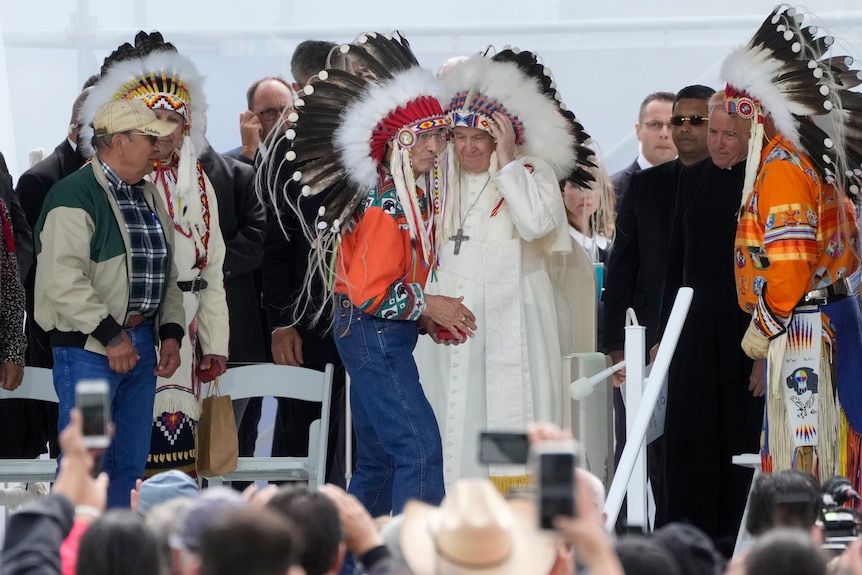 A man in traditional dress stands partly in front of Pope Francis who is wearing robes and a traditional Indigenous head piece. 