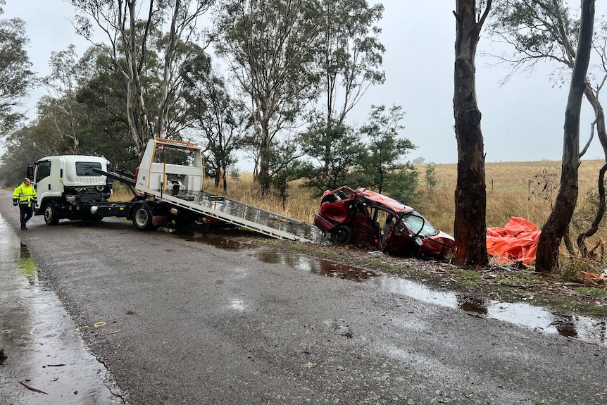 A red car extensively damaged in a crash is loaded onto a truck on a rainy day.