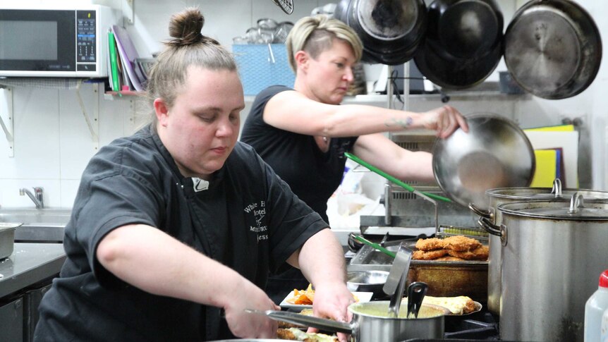 Two women dressed in black work in a commercial kitchen, preparing schnitzels, surrounded by steel pans and pots.
