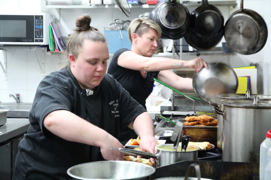 Two women dressed in black work in a commercial kitchen, preparing schnitzels, surrounded by steel pans and pots.