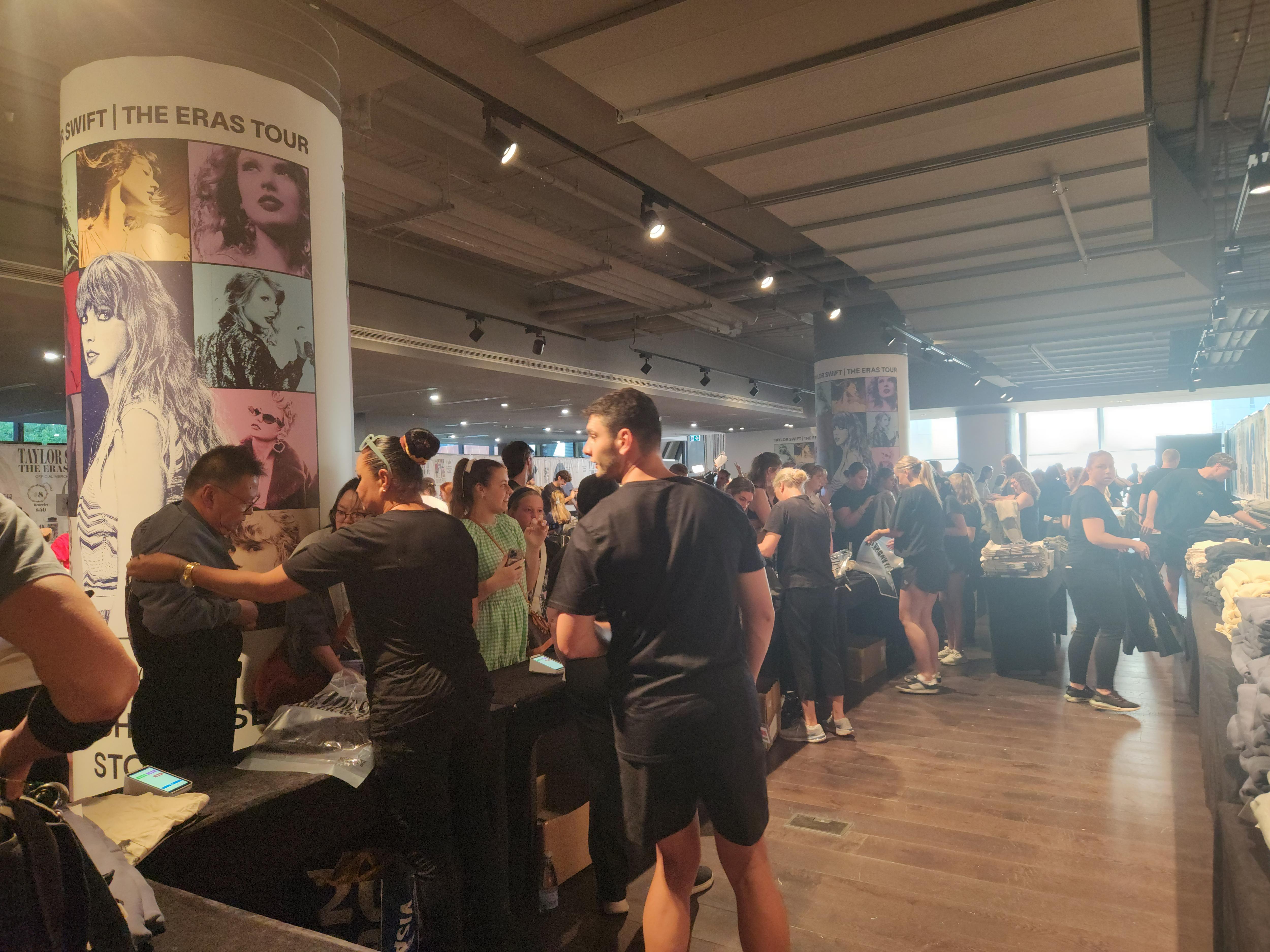 A crowd of people standing by tables full of Taylor Swift merchandise with posters of her on poles.