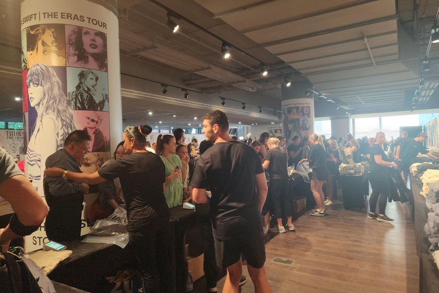 A crowd of people standing by tables full of Taylor Swift merchandise with posters of her on poles.