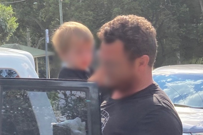 A man wearing a black t-shirt carries a small boy a car. Both their faces are blurred.