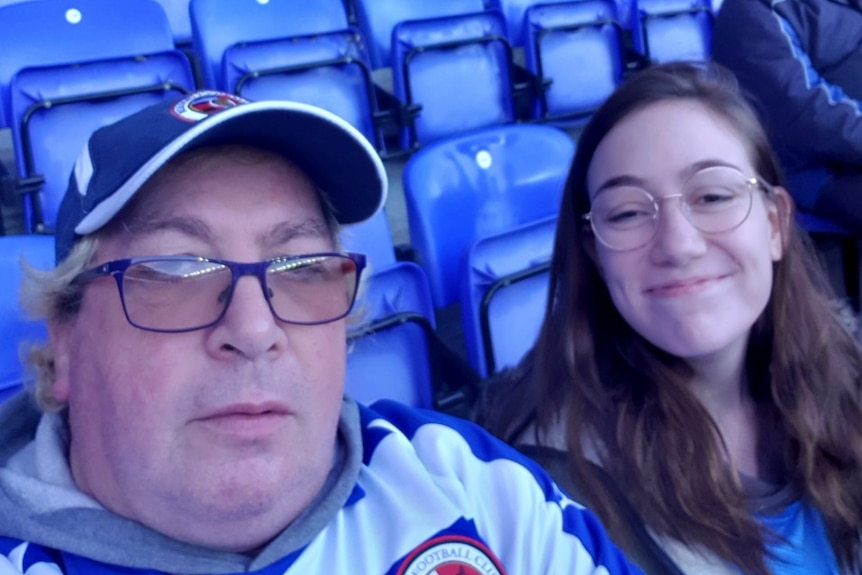 A selfie is taken of a Caucasian man and his daughter who are seated on bright blue stadium seats.