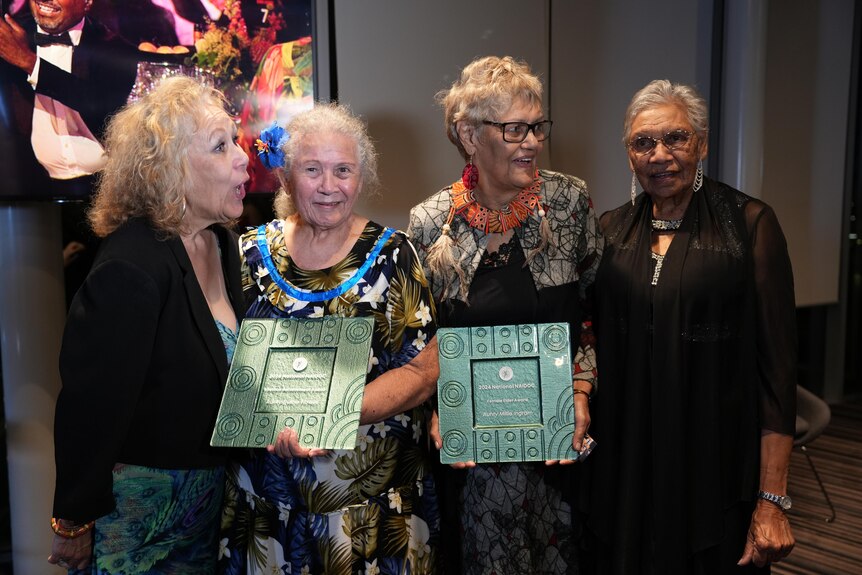 Four older Indigenous women pose with award plaques as they laugh and chat.
