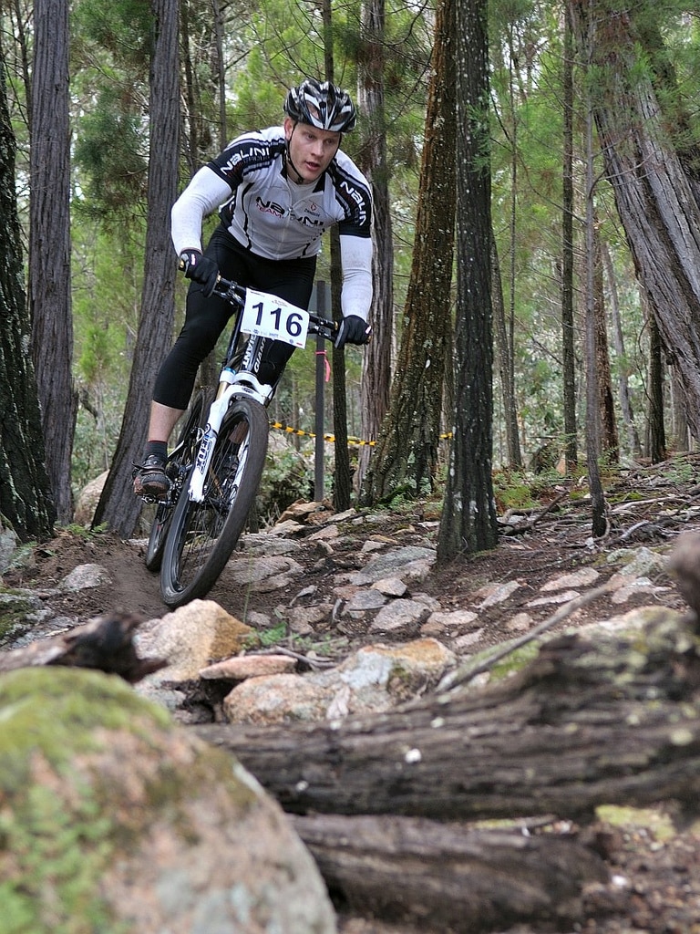 A man with the number 116 on his bike riding over rocky terrain, surrounded by trees.