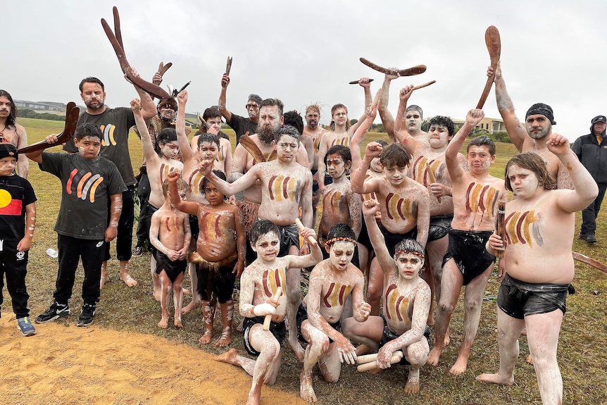 A group of people pose for a photo, some are holding boomerangs and sticks and wearing face paint.