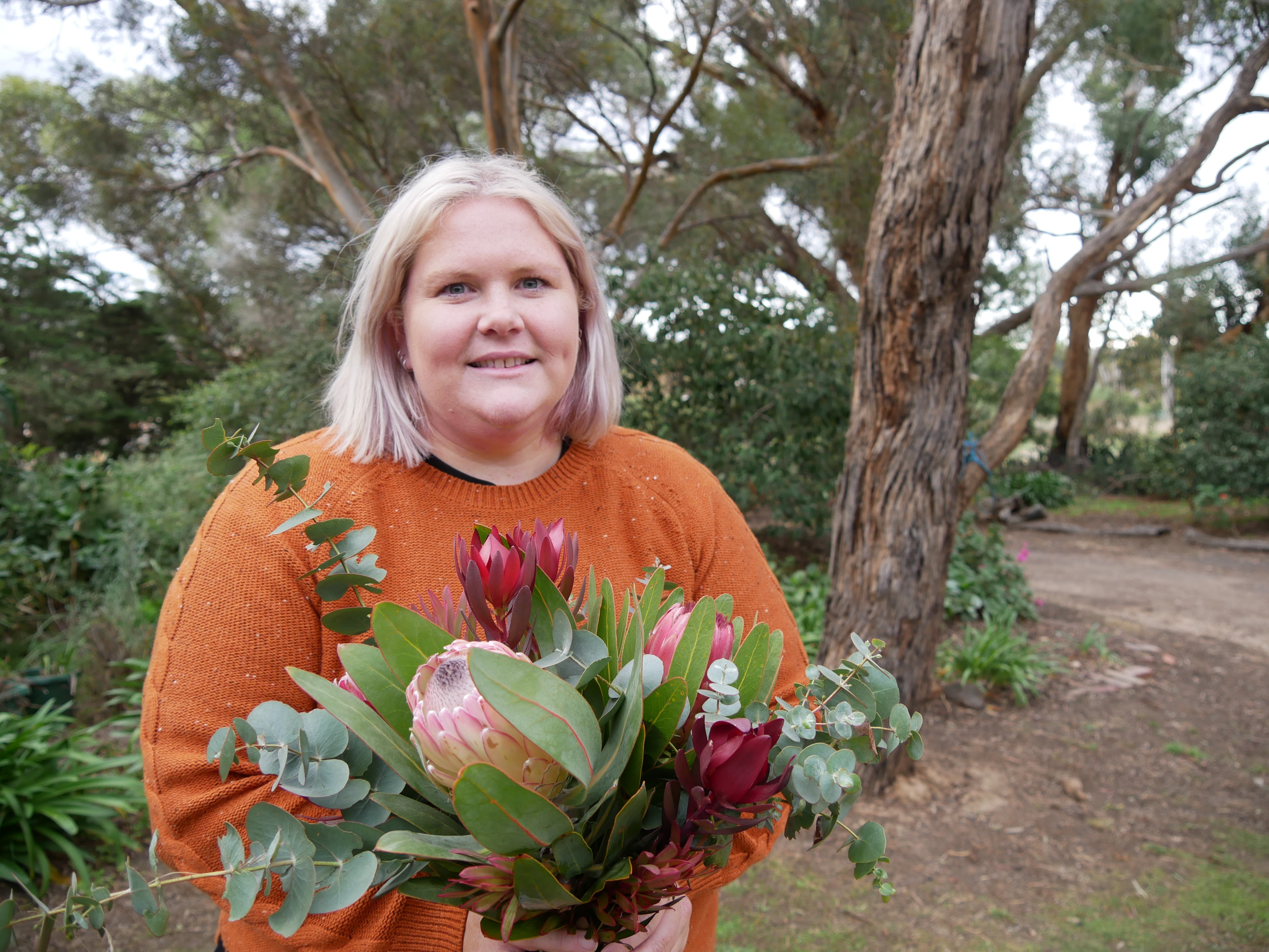 Uma mulher loira de suéter segura um buquê de flores em pé perto de uma árvore.