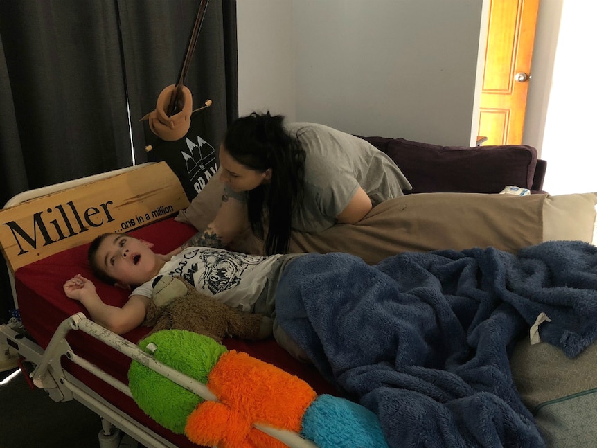A mum leaning over her eight year old son in a hospital bed at home.