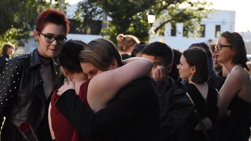 A number of young people mingle and talk in the background as two hug.