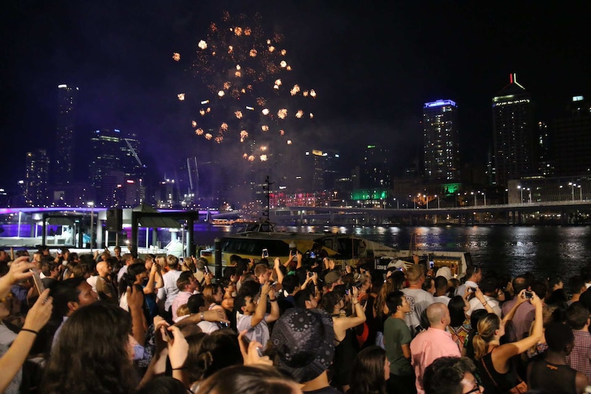 A large crowd of people standing next to the Brisbane River take photos of fireworks in the sky.