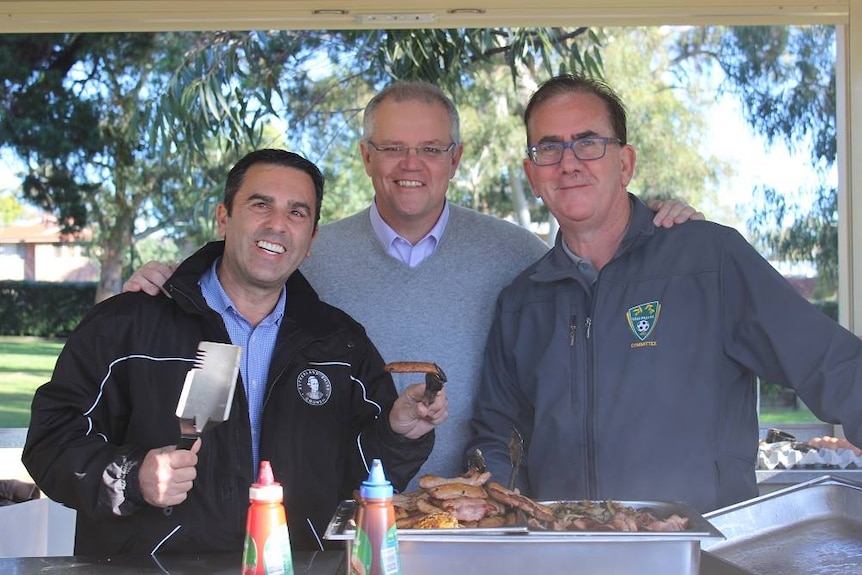 Three men stand around a barbecue and smile at the camera