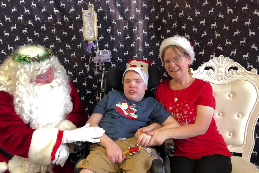 A woman in a red shirt and Christmas hat next to her son who is in a wheelchair meet Santa